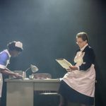 Two women at work, wearing 1960s blue nurses uniforms. The woman on the right is sat on the edge of a desk, reading some papers.