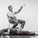 Kobna Holdbrook-Smith and Paapa Essiedu performing a scene in the rehearsal room. Paapa is lying on the floor and Kobna is sitting in a chair gesturing to the right.