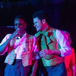 Two actors on stage laughing and sharing a moment, one holding a drink, both in colorful period waistcoats and white shirts