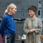 Two women having a conversation. The woman on the left is wearing a blue jumper with her hands behind her back. The woman on the right is wearing a leopard print dress and her hands are clasped in front of her.