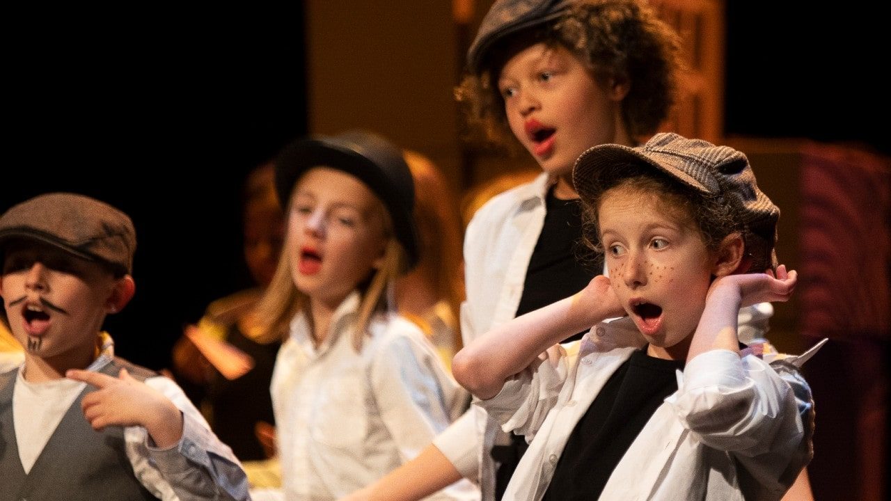 Four children onstage performing in a play, wearing flat caps and with painted freckles and a moustache.