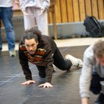 A man with brown curly hair, tied back, crawling on the floor. He is wearing a brown jumper with colourful stripes on the front. Beside him other cast members are also crawling.