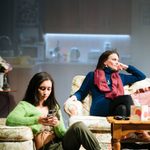 Yasmin sits on a floral patterned green rug, with a boarder, she has her arm up resting on a floral patterned footstool. She is wearing a green cardigan, a brown crop top khaki coloured combat trousers and white trainers she has long brown hair. She is looking down at the mobile phone she is holding in her hands. Behind her on a sofa sits Liz a woman with a brown long bob, She wears a Royal Blue roll neck jumper dress with a vertical striped pattern, over the jumper she wears a red cotton scarf, a silver chain what has a silver ring at the end and silver hoops. She is looking up, with her head resting on her hand. Her elbow is resting on the back of the sofa.