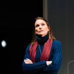 A white woman with a brown long bob stands with her arms folded. She wears a Royal Blue roll neck jumper with a vertical striped pattern, over the jumper she wears a red cotton scarf, a silver chain and hoops. She is looking up.