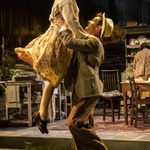 Tom Riley as Gerry lifting Louisa Harland as Agnes as a part of a dance. Tom wears a suit and hat and Louisa wears a cream floral dress.