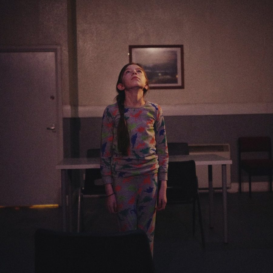 A young girl stands straight, in a dimly lit functional room, with a doorway off to the right open onto an illuminated space.