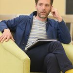 A young man with brown hair and a short beard sits in a chair, with one hand holding his ear. He is wearing a blue and white striped T-shirt, a blue jacket, blue trousers and yellow socks.