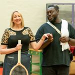 A woman with a blonde bobbed hair cut stands, shes holding a old fashioned tennis racket. She is wearing a black t-shirt with a leopard motif, she wears a denim skirt. She is holding the hand of a man with a light black bears and hair back in a short ponytail. He is looking at her adoringly, he has a white shawl over one shoulder.