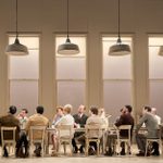 Mark Gatiss is gesturing with his hand. He wears a brown-green corduroy suit, white shirt, black belt and brown shoes. A group of people sit around a long table to the right of the man, listening to him intently.