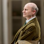 A close-up of Mark Gatiss. He wears a brown-green corduroy suit jacket, white shirt and dark green tie.