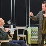 A young man with dark blonde hair in a tweed coat and green shirt stands gesturing towards a balding man sitting down. The balding man is listening with hands clasped, whilst wearing a white shirt and fair isle vest.