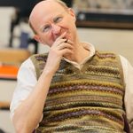 A balding man in a white shirt and a fair isle sweater vests sits in a chair. A book is in his lap. He wears glasses and holds a pen and listens to someone off camera with intent.