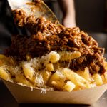 A tray of chips covered in parmesan and with a beef in gravy topping being piled on top.