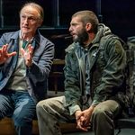 Two male characters, one using his hands to emphasise a point, the other, with a large military style rucksack,listening. Both sitting next to each other on a bench.