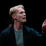 A man in a light T-shirt and dark jacket is mid-speech, pointing his finger.