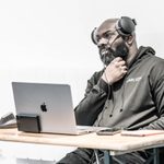 Mikey J Asante sitting in the rehearsal room with a laptop and headphones on. He is wearing a black hoodie.