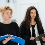 Milly Alcock and Nia Towle stad looking off to the side, both holding their scripts open.