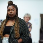 Nadine Higgin stands in a white space with blurred figures in the background, looking into camera.