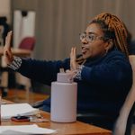 A woman with ginger box braids, wearing patterned glasses and a dark blue jumper. She is gesturing with her hands with one hand extended and the other close to her chest. There is a script and purple water bottle on the table in front of her.