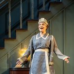 A woman in 1930s gray maid's uniform stands in front of a teal staircase.