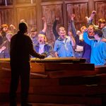 A group of men in blue, black and grey clothing stand behind a wooden fence in an enclosure of sorts. They all raise their fists and shout in rebellion. A man on the other side faces them.