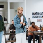 Nick Fletcher, Caitlin FitzGerald, and cast members standing and sitting in a large white space, with wooden chairs and table, acting out a scene.