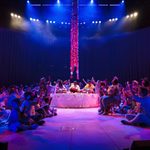 A group of drummers sat in a central table with a plume of red string hanging from the ceiling, surrounded by the community cast.