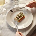 Eel strips on a confit potato, with green micro herb garnish. Sits on a white plate with a black and white logo. Hand on either side of the plat hold a knife and fork aloft on either side. There is a glass of white wine to the left and a wine bottle is just in shot.