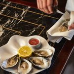 A plate of oysters in a special white oyster dish sits on a black marble bar. There is a lemon in the middle. To the side out of shot apart from his hands which are mid way through shucking another oyster.