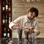 A bartender is stood at the bar, he has floppy dark hair and is earing a cream service jacket. In front of him is a cocktail shaker. He is pouring into a shot glass. Behind him is a wall of concrete, to the left is a stack of shelves with various bottles of alcohol. Above his head are rows of glasses hanging from the bar.