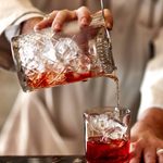 A cocktail is mid way through being made. A glass jug filled with ice ice pouring into a glass tumbler. The liquid is light red.