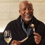 An older man with a grey beard is sitting eating a dish he is looking off to the side and is smiling, in front of him is a glass of wine and a saffron bun.