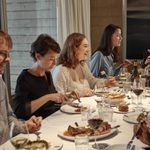 A group of people of various ages, sat at a long table, on the table is a white table cloth, the table has various dishes along the length. There is a painting at the end hanging on the wall. Everyone is in conversation.