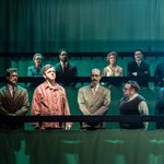 Nine people in suits with hospital curtains lowered to recreate the benches in the House of Commons. Michael Sheen, in red striped pyjamas, is stood among them.