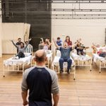 A man looks at five people sitting on beds with their arms raised.