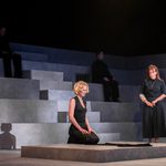 Desdemona kneeling and Emilia standing, both pensive, centre stage with figueres seated above and around them.