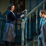 A woman in navy blue 1930s dress and a man in a blue three piece suit look at each other in front of a teal staircase.