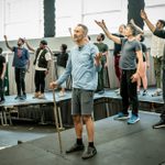 A man wearing a blue long-sleeved T-shirt and grey shorts stands with a walking stick in hand. Behind him are a group of men and women with their arms stretched out.