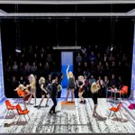 Seven blond-haired women and one man are dancing on a stage, set with orange chairs, with an audience behind them.