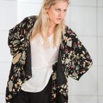 A woman wearing a black floral patterned shirt, open over a white t-shirt, stands, leaning forward slightly. A white tiled wall is behind her.