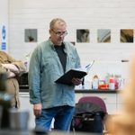 Two men in rehearsals. The man on the left is in a brown shirt with his arms folded and a serious expression. The man on the right is wearing a blue shirt and glasses, holding a script in his left hand.