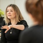 Rachel O'Riordan wearing a dark top, arm raised, in the rehearsal room, with th eback of someone's head in the foreground.