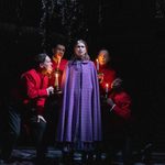 A tense stage scene with four actors in red holding candles around an actress in a purple dress, their faces illuminated by the candlelight, creating a dramatic atmosphere.