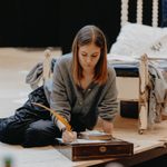 A woman with brown hair sat on the floor at the end of a bed. She is writing with a yellow quill on some paper, using a model-antique writing slope.