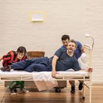 Michael Sheen lies on a bed while two men stand behind the bed and push it forward.