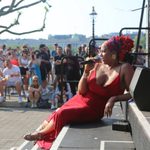 A performer wearing a long red dress with an elaborate colourful headscarf and large gold earrings. She is sat on the steps of a stage at an outdoor summer event, holding a microphone. A crowd of people are watching from the side.