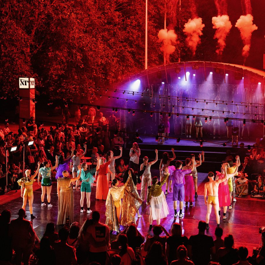 A large group of performers in colourful and vibrant outfits performing at an outdoor summer event in the evening. They are surrounded by a large crowd watching.