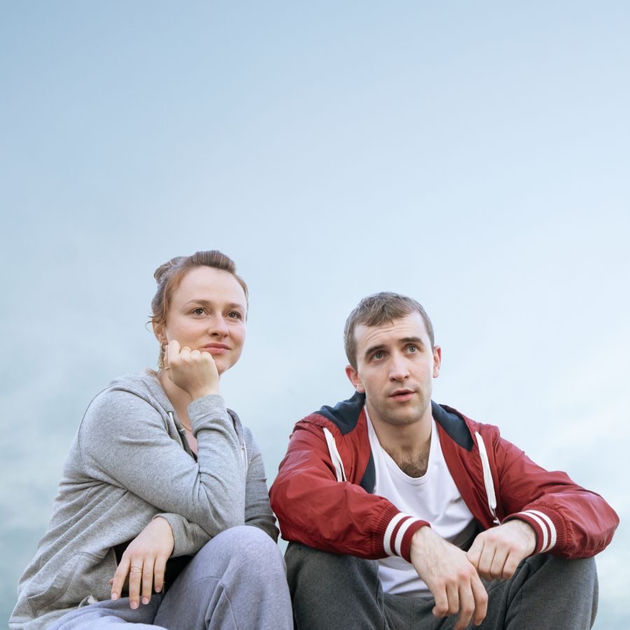 Romeo and Julie poster photo of Callum Scott Howells and Rosie Sheehy, wearing tracksuit tops and bottoms, sat side-by side against a hazy, blue sky