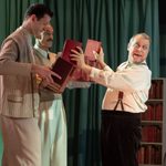 A man in a white shirt and suspenders holding two red books. Two men hold rows of books that the other books are taken from.