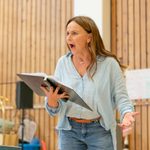 A woman stands in a light blue linen shirt, she is wearing jeans. She is holding a black folder in front of her. She has light brown long hair that is lightly curled, her mouth is open in a shocked and concerned expression